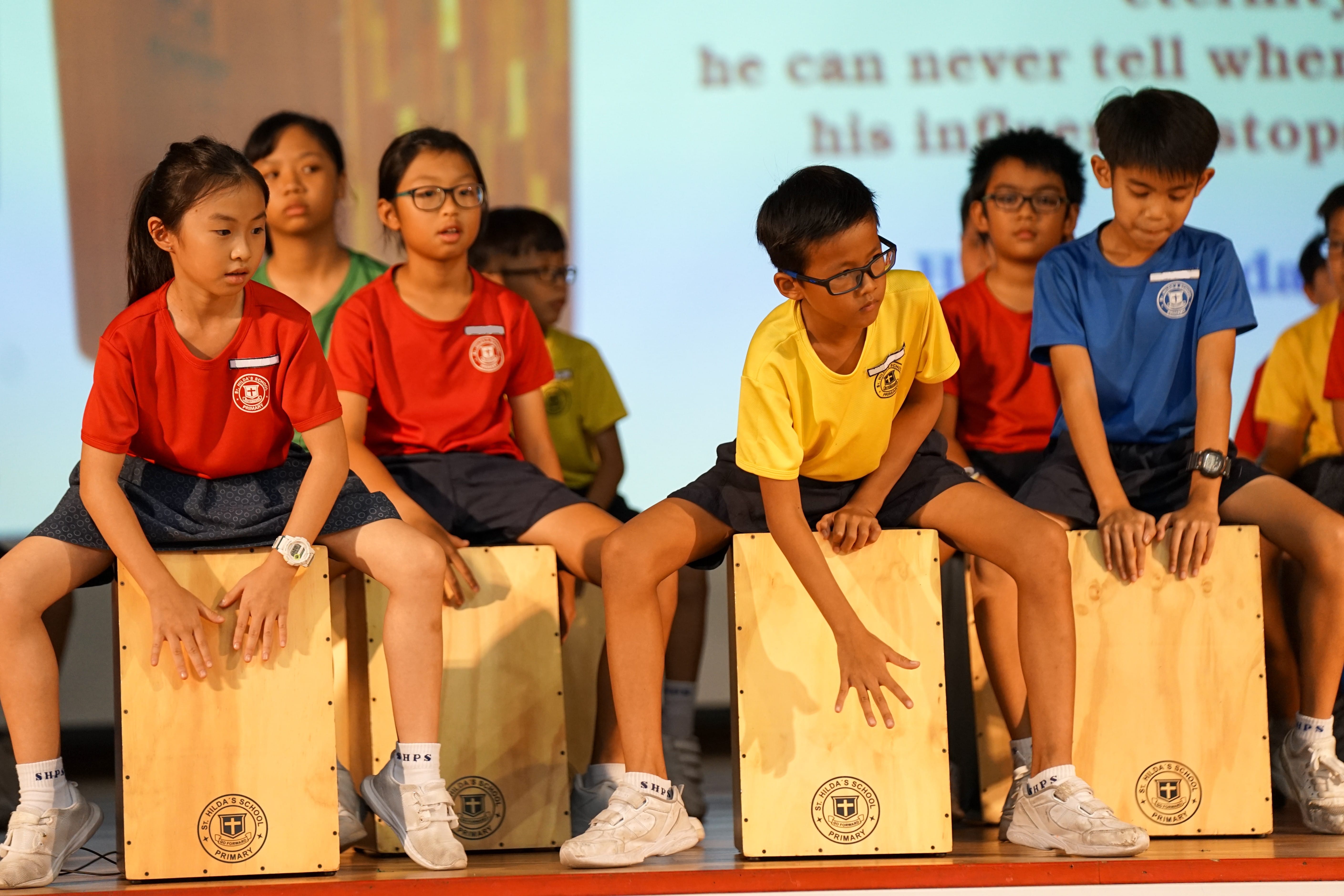 Students are performing a musical routine by sitting on and striking wooden box drums, known as cajóns, with their hands.